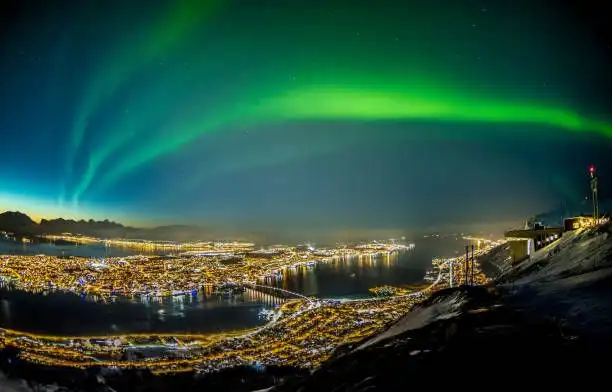 Las auroras boreales verdes se extienden por el cielo nocturno sobre una ciudad costera brillantemente iluminada rodeada de agua y montañas nevadas.