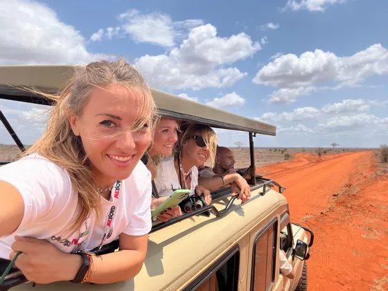 Un gruppo WeRoad scatta un selfie da un veicolo safari scoperto su una strada sterrata rossa nella savana.