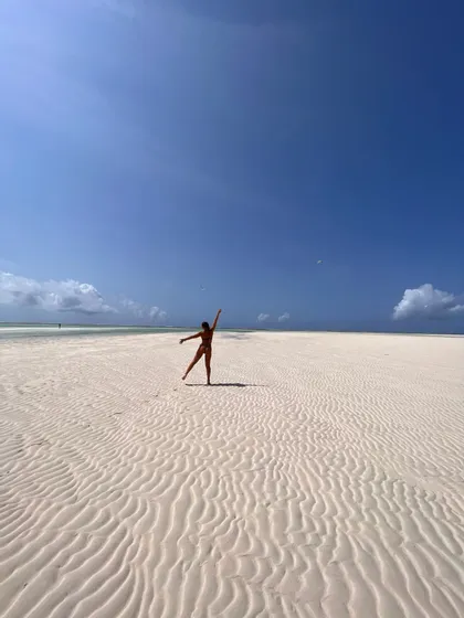 Una donna in bikini si trova su un'ampia spiaggia con sabbia bianca increspata, in posa con le braccia distese sotto un cielo azzurro.
