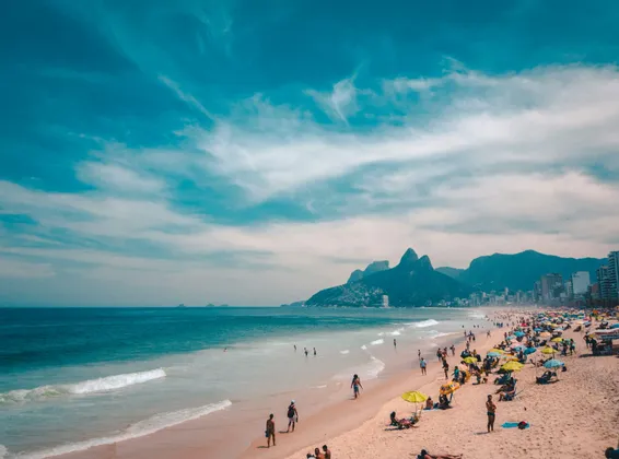 Une plage de sable animée, avec des baigneurs et des personnes prenant le soleil, une ville et des montagnes en toile de fond sous un ciel bleu éclatant.