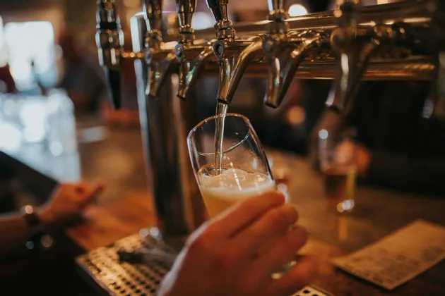 Primo piano di una birra alla spina versata da un rubinetto cromato in un bicchiere inclinato tenuto da una mano al bar.