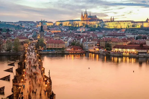 Un ponte di pietra illuminato e affollato di persone su un fiume, con una città storica e un castello su una collina al tramonto.