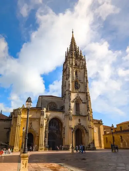 Una ornamentada catedral gótica con una alta aguja se alza en una plaza de piedra bajo un cielo azul con nubes blancas.