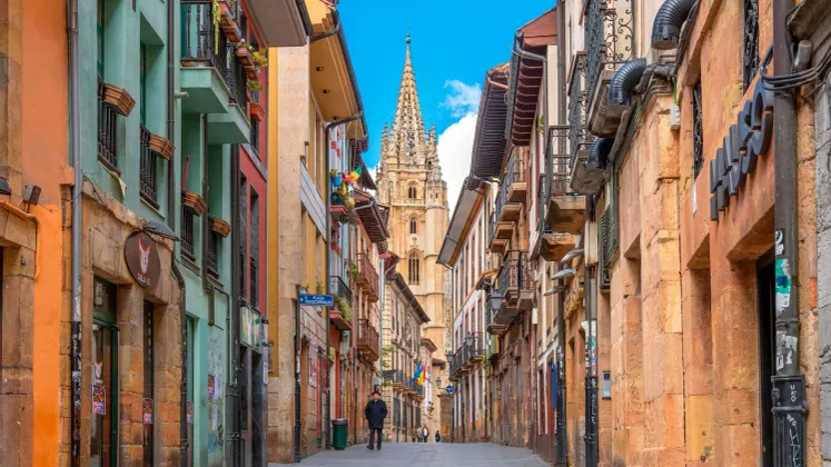 Vista de una calle estrecha con edificios coloridos, que conduce a una alta y ornamentada aguja de catedral bajo un cielo azul.