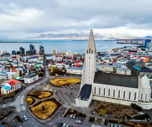 Una vista aérea de una gran iglesia moderna con vistas a una colorida ciudad costera con montañas nevadas en la distancia.