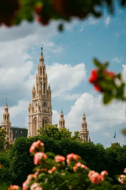 Una torre dell'orologio in stile gotico si erge sopra alberi verdi, vista attraverso fiori sfocati sotto un cielo parzialmente nuvoloso.