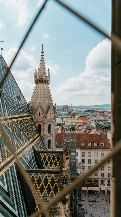 Vista attraverso una recinzione del tetto piastrellato colorato e della guglia di una cattedrale, con vista su un paesaggio urbano europeo sotto un cielo parzialmente nuvoloso.