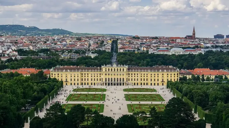 Una vista sopraelevata di un grande palazzo giallo con giardini all'italiana, sullo sfondo un'estesa città e colline.