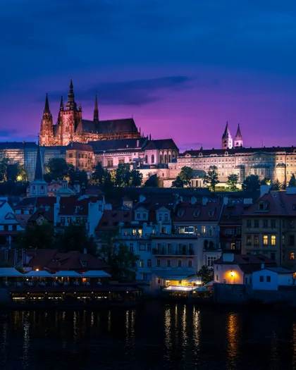 Un castello e una cattedrale illuminati su una collina che domina gli edifici della città e un fiume al crepuscolo sotto un cielo viola