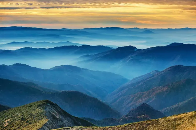 Blaue Gebirgsketten ziehen sich in die Ferne, mit nebelgefüllten Tälern unter einem goldenen Sonnenuntergangshimmel.