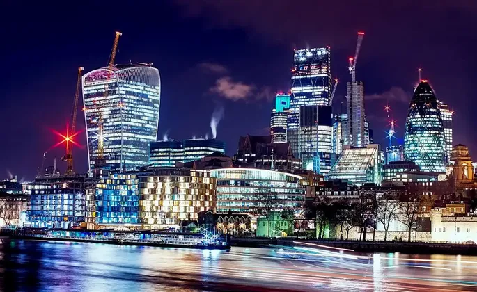 Uno skyline urbano di grattacieli ed edifici illuminati di notte, visto dall'altra sponda di un fiume con scie luminose colorate sull'acqua.