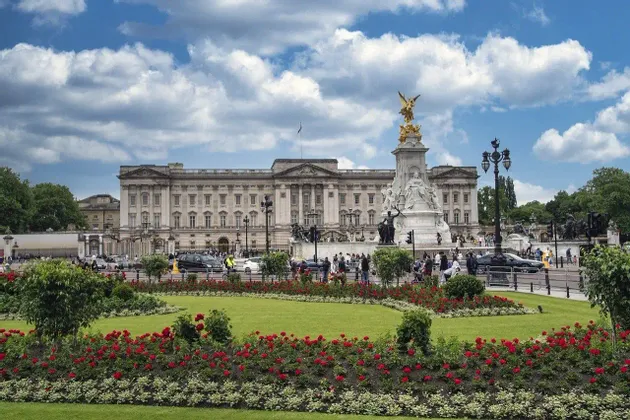 Un grande palazzo neoclassico e una statua commemorativa visti da un giardino con rose rosse, sotto un cielo parzialmente nuvoloso.