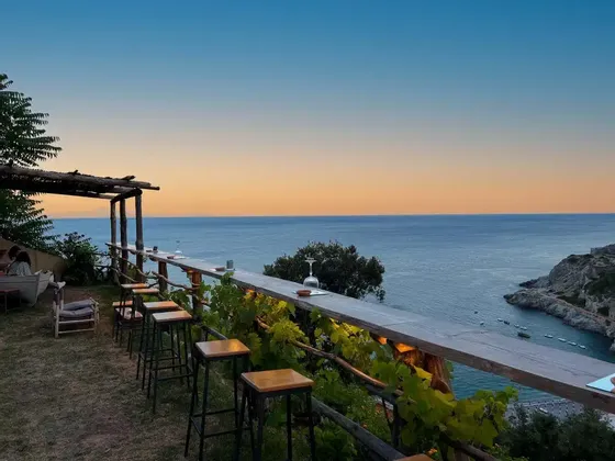 Un bar all'aperto con un lungo bancone in legno e sgabelli si affaccia sul mare e su una costa rocciosa al tramonto.