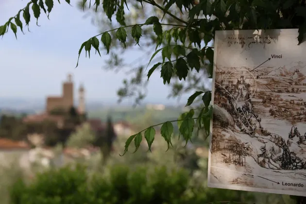 Una mappa di Vinci, opera di Leonardo, pende da un ramo d'albero, con il paese visibile sullo sfondo sfocato.