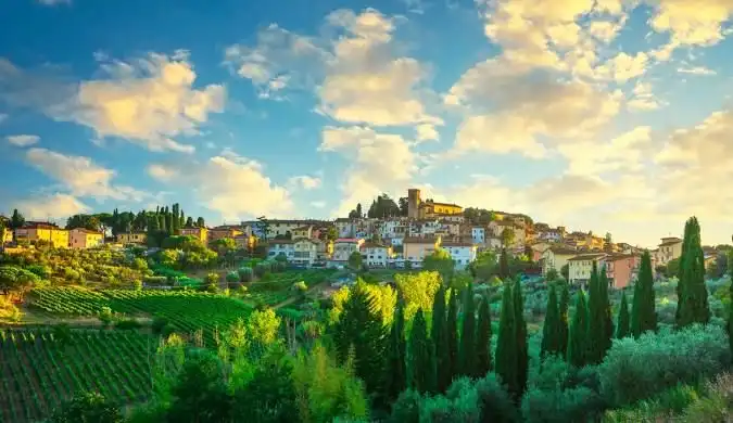 Un borgo illuminato dal sole su una collina circondata da verdi vigneti e cipressi sotto un cielo azzurro con nuvole sparse.