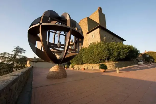 Una grande scultura sferica di metallo si erge su un piedistallo di mattoni su una terrazza pavimentata accanto a un edificio storico in pietra sotto un cielo sereno.