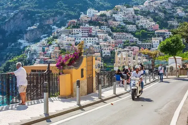 Una persona en una scooter blanca conduce por una carretera costera con vistas a un pueblo en la ladera con casas coloridas y vegetación exuberante.