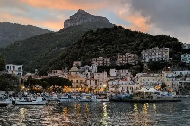 Un pueblo costero con su puerto lleno de barcos, iluminado al anochecer con una gran montaña arbolada al fondo.