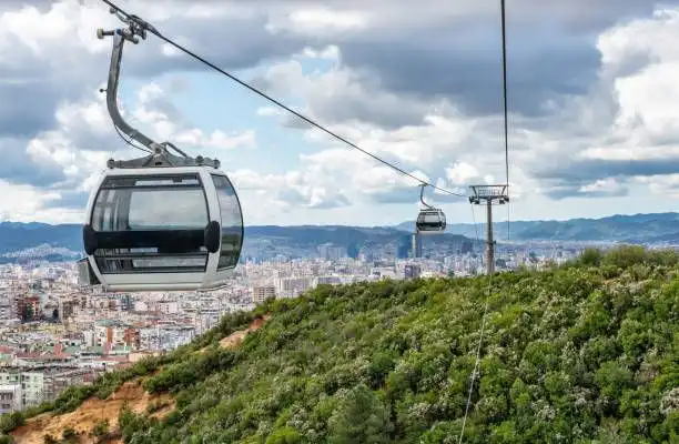 Una moderna gondola di funivia si muove lungo una linea sopra una verde collina, con una città estesa visibile sullo sfondo.