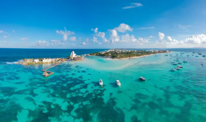 Vista aerea di una penisola tropicale con resort e barche ancorate in acqua cristallina e turchese sotto un cielo blu con nuvole.