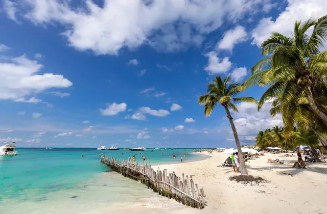Una spiaggia tropicale soleggiata con palme, sabbia bianca e acqua turchese. Le persone si stanno rilassando e nuotando, con barche ancorate al largo.