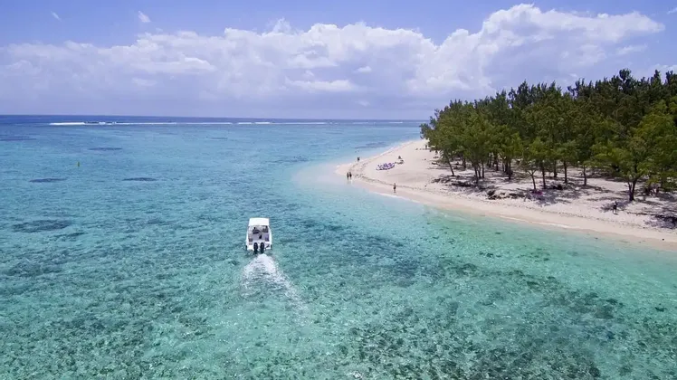 Vue aérienne d'un bateau à moteur blanc naviguant sur des eaux turquoises claires vers une plage de sable bordée d'arbres.