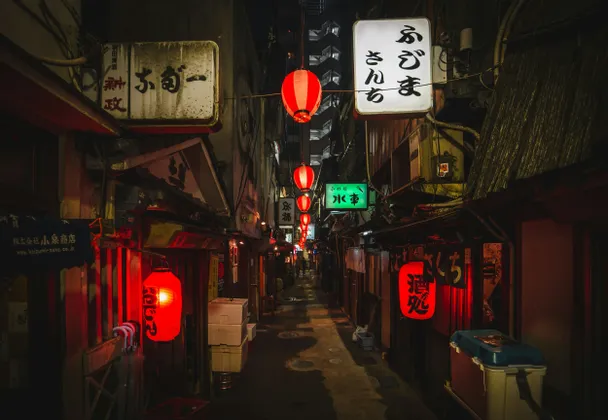 Un vicolo urbano stretto di notte, illuminato da lanterne di carta rosse luminose e insegne con caratteri giapponesi.