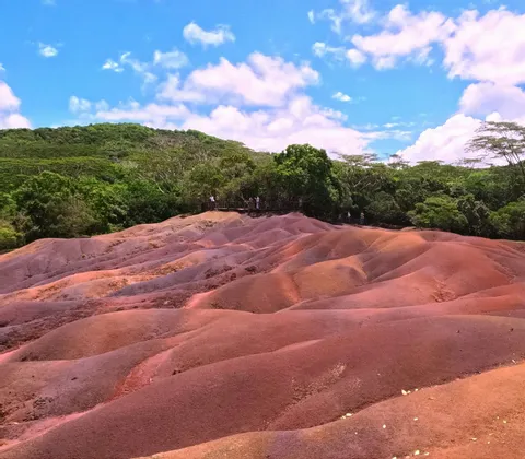 Des dunes ondulantes de terre multicolore aux nuances de rouge et de brun se détachent sur un fond de forêt verdoyante sous un ciel bleu parsemé de nuages.