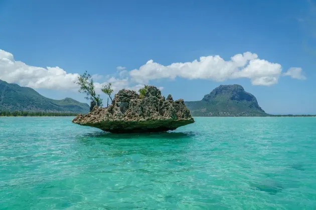 Une grande formation rocheuse avec de petits arbres se trouve dans une eau turquoise claire, avec des montagnes en arrière-plan.