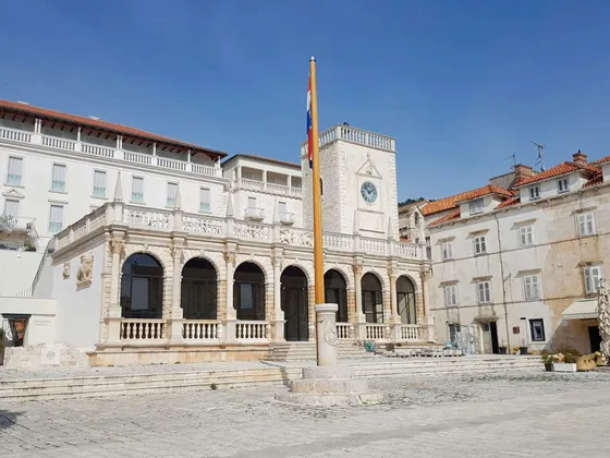 Un edificio storico in pietra con una torre dell'orologio e una galleria ad arco si erge in un'ampia piazza lastricata accanto a un'alta asta della bandiera.