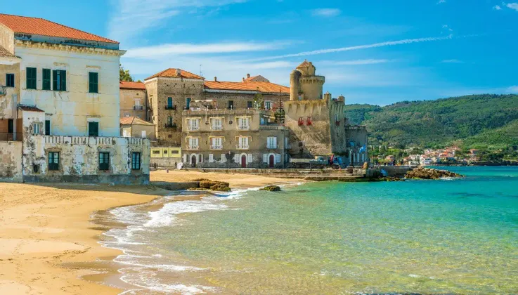 Una spiaggia di sabbia con acque cristalline turchesi di fronte a una cittadina costiera con una fortezza in pietra e colline verdi sullo sfondo.