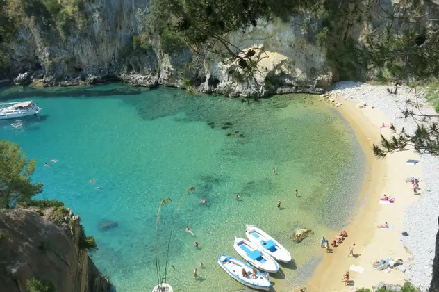 Una vista dall'alto di una caletta appartata con acqua turchese, dove le persone nuotano, prendono il sole su una spiaggia sabbiosa e si rilassano su piccole imbarcazioni.