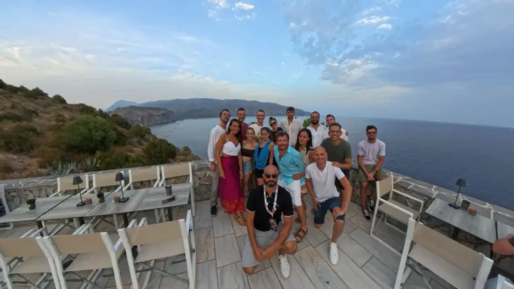 Un gruppo WeRoad in posa per una foto su una terrazza di pietra con vista su una baia e una costa montuosa.