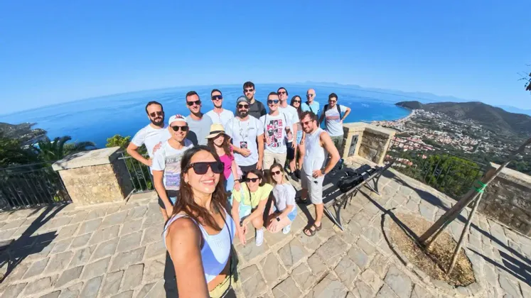 Un viaggio di gruppo WeRoad si scatta un selfie grandangolare su un punto panoramico che si affaccia su una città costiera e il mare blu.