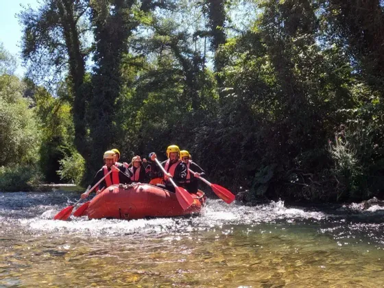 Un viaggio di gruppo WeRoad pagaia su un gommone rosso lungo un fiume, circondato da una fitta foresta verde sotto un cielo soleggiato.