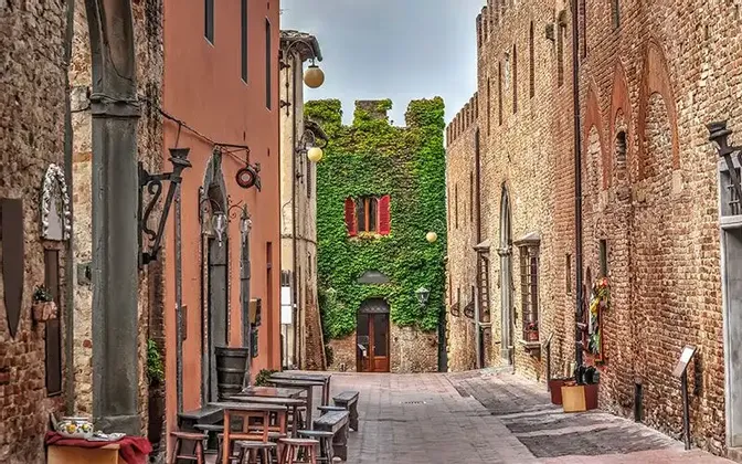 Uno stretto vicolo in mattoni con tavoli da caffè all'aperto conduce a un edificio storico coperto di edera verde.