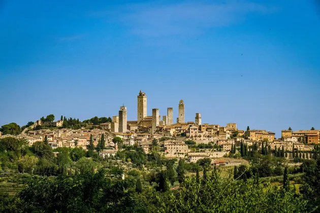 Una città storica con molteplici torri in pietra sorge su una collina verde lussureggiante, circondata da alberi sotto un cielo azzurro limpido.