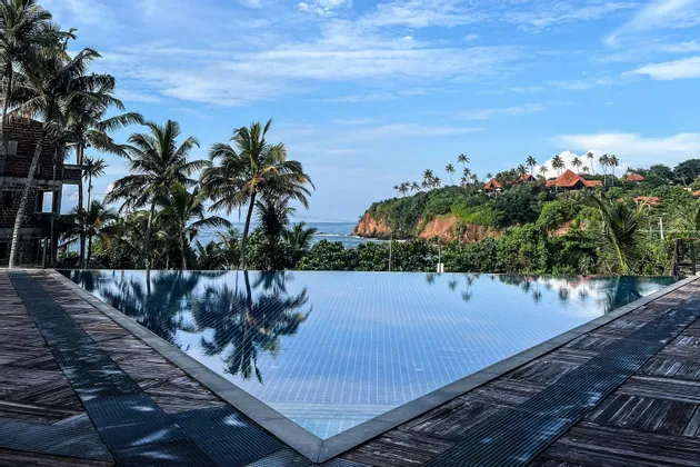 Una piscina a sfioro con un ponte in legno si affaccia su una costa tropicale con palme, una scogliera verde e l'oceano.