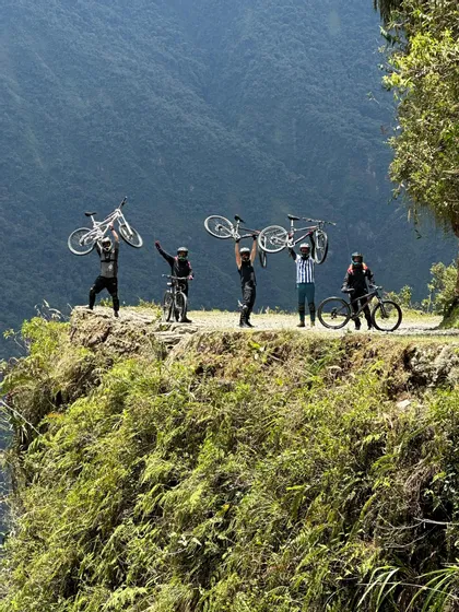 Un viaje en grupo de WeRoad con ciclistas levantando sus bicicletas de montaña en el borde de un acantilado con un exuberante paisaje montañoso de fondo.