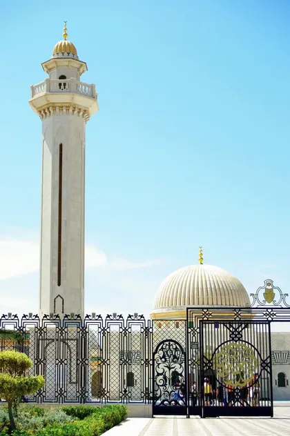 Un minareto bianco e la cupola di una moschea si ergono dietro un cancello di metallo ornato sotto un cielo blu brillante.