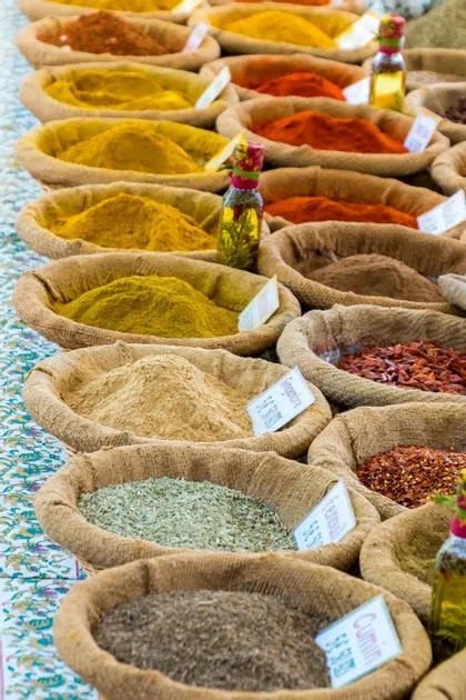 Spezie macinate ed erbe intere colorate, esposte in cesti di juta su una tovaglia a motivi, al mercato.