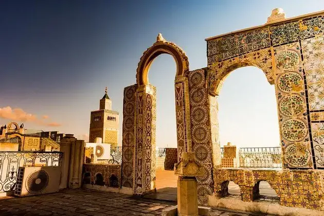 Archi piastrellati decorati su una terrazza sul tetto si affacciano su uno skyline cittadino con un minareto al tramonto.