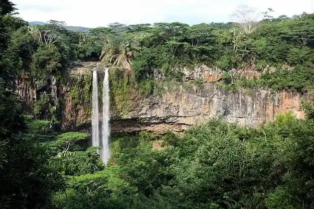 Ein hoher Wasserfall stürzt eine felsige Klippe hinab, umgeben von dichtem, grünem Dschungel unter einem hellen Himmel.