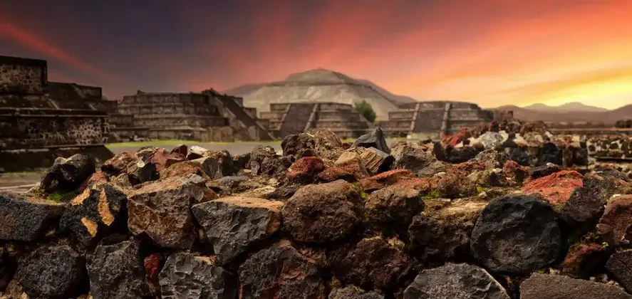 Antiche piramidi di pietra si ergono dietro un primo piano di aspre rocce vulcaniche sotto un tramonto vibrante.