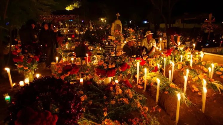 Un cimitero affollato di notte, con tombe adornate da abbondanti fiori e illuminate da numerose candele alte e accese.