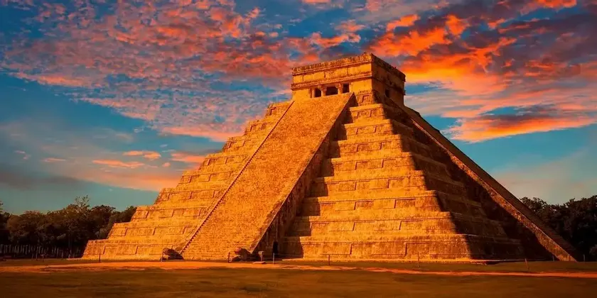 Una grande piramide a gradoni in pietra è illuminata dal sole al tramonto, contro un cielo blu con nuvole arancioni drammatiche.
