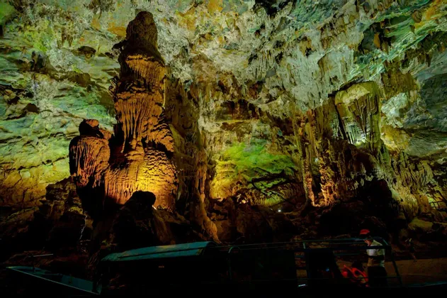 Una barca con un gruppo WeRoad naviga attraverso un'immensa caverna, con stalattiti e stalagmiti illuminate da luci verdi e gialle.
