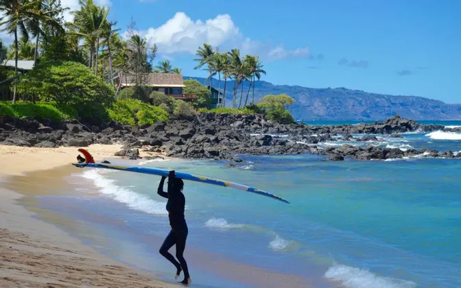Un surfista con muta nera porta una longboard lungo una spiaggia tropicale con acqua turchese.