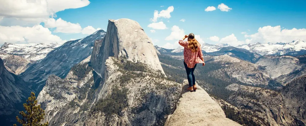 Una donna con una camicia a quadri si erge su una cima rocciosa, ammirando una vasta catena montuosa con cime innevate sotto un cielo azzurro.