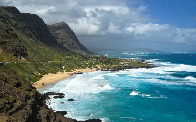 Una costa verde e frastagliata con una spiaggia sabbiosa incontra l'oceano turchese con onde bianche sotto un cielo nuvoloso.